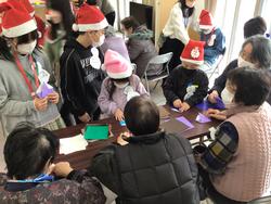 サンタ帽子をかぶった子どもたちが高齢者と一緒にテーブルを囲み、折り紙を使って工作をしている様子を写した写真