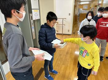 廊下で2人の男子児童が紙製の箱を持って並んで立ち、募金箱にお金を入れている黄色いトレーナーを着た男児の後ろに、列になって並んだ児童たちが募金の順番を待っている様子の写真