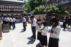 奥に提灯が並ぶ拝殿や玉垣が見える神社の境内で、手を合わせて参拝している制服姿の生徒たちの写真