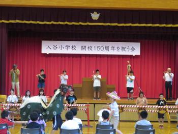 舞台には「入谷小学校 開講150周年を祝う会」の幕が飾られ、舞台上で演奏する子供達、舞台の下で獅子舞が踊っている様子を写した写真