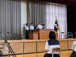 体育館の舞台上で制服姿の生徒数人が演台の後ろに立ち、前方の椅子に別の生徒たちが座っている始業式の様子を後方から撮影した写真