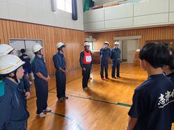 体育館でヘルメットと作業着を着用した消防職員の方々が横一列に並び、中央あたりに立っている赤いビブスをつけた職員の方が話をし、向かい合って立った生徒たちが注目している様子の写真