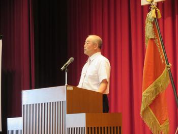 舞台の演台に立ちマイクを使い話をしている男性を右斜め前から写した写真