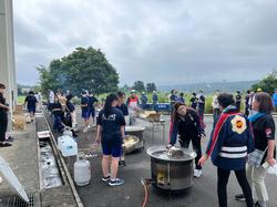 建物横の屋外スペースで大勢の生徒や町の職員などが集まり大きな鍋で料理を作りながら準備や交流を行っている写真