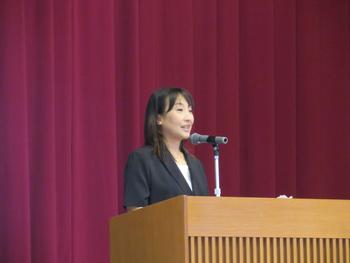 舞台の演台に立ちマイクを使い話をしている女性を左斜め前から写した写真