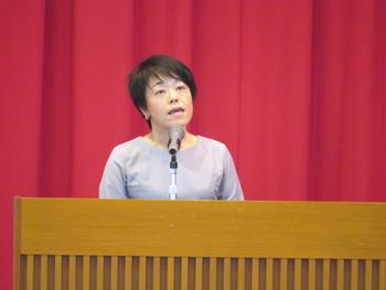 舞台の演台に立ちマイクを使い話をしている女性を正面から写した写真