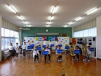 楽器を持つ十数名の吹奏楽部員たちが、白いシャツに青いスカーフをつけ、広い部屋で横に広がって並んでいる演奏風景の写真