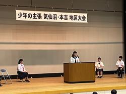 壇上に掲げられた「少年の主張 気仙沼・本吉 地区大会」と書かれた横断幕の下で、中央の演台に立って発表する女子生徒と、後ろで椅子に座って話を聞いている複数の生徒が写った写真