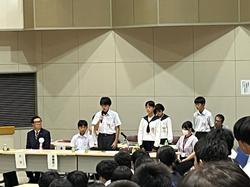 席に着いた生徒たちが注目する中、長机の席の生徒のうち数名が立ち、その中の1人の男子生徒がマイクを持って発表をしている写真