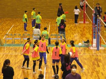 体育館で行われているバレーボールの試合で、黄色Tシャツにピンク色のビブスを着た女子チームがコートの中で円になっている様子を捉えた写真