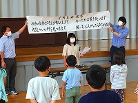 体育館の舞台の前で2人の大人が横断幕を広げ、中央に立つ人が紙を読み上げ、複数の児童が前を向いて話を聞いている学校行事の様子の写真
