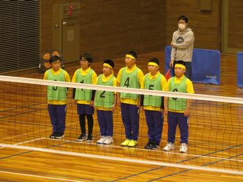 体育館のバレーボールネット前に、黄色いTシャツに、背番号が付いた黄緑色のビブスを着用した6人の男子児童たちが並んでいる様子の写真