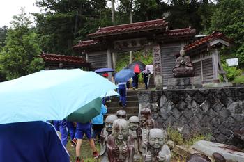 児童達が傘をさし列をなしながら、お地蔵さんが並ぶ道を通り、階段の先の立派な門の中に進んでいる写真