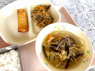 ピンクのトレーに給食が左から時計回りにはるまき、マーボーなす、中華きのこスープ、ごはんと並んでいる写真