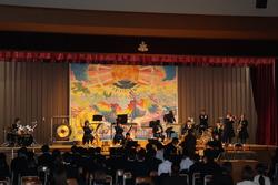 ステージの上で吹奏楽部の生徒たちが様々な楽器を演奏し、客席に座ったたくさんの生徒たちが演奏を聴いている様子の写真