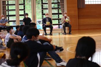 体育館の入り口側にパイプ椅子に座った男性が4名並び、体育館内では児童が整列して床に座っている写真