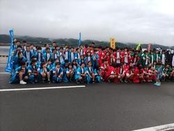 曇り空の下、水色、赤、緑などの法被を着た大勢の生徒たちが、色ごとに並んで、後ろに立ったのぼり旗と一緒に記念撮影をしている写真