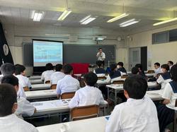 教室にたくさん並んだ長机に2人ずつ座った生徒たちが、前方の黒板前のスクリーンと、横で話をしている男性の方を向いている写真