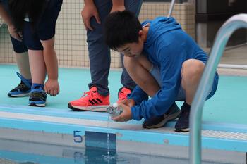 一人の男子児童がプールサイドにしゃがみ、手に持った容器からプールに水を注いでいる写真