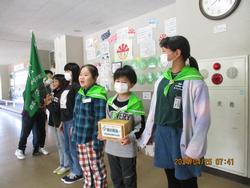 廊下にバンダナを肩に付けた児童が募金箱を持ち横一列に並んでいる写真