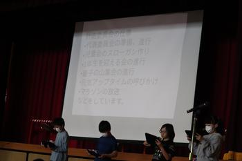 舞台上のスクリーンに文字が映し出され、舞台の下に4名の児童が1列に並んでタブレット端末を使いながら活動紹介を行っている写真