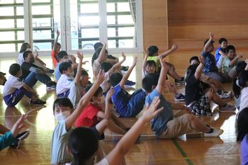 体育館の床に座った児童たちの多くが右手を上にあげて挙手をしている様子を横から写した写真
