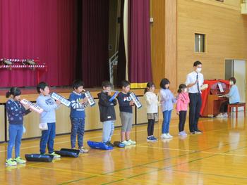 舞台の前で鍵盤ハーモニカやカスタネットを1列に並んで演奏している8名の児童と先生2名の写真