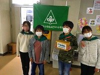 マスクをつけた小学生4人が、緑色の旗の前で募金箱を手に持ち、カメラに向かって立っている様子の写真