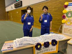 広い会場に置かれた「みやぎ心のケアセンター」の案内が貼られ、たくさんの資料が広げられた長机の後ろに立ったジャージ姿の男女の生徒がピースサインをしている写真