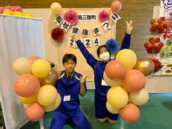 「南三陸町 福祉健康祭り2024」の文字と花飾りなどが飾られた壁の前で、左右に置かれた黄色やオレンジの風船の束の間に座ったジャージ姿の男女の生徒がピースサインをしている写真