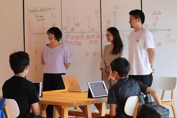 タブレットを目の前に置いた男の子2名が前方に並んでいる女性2名、男性1名のほうを向いて机に座っている様子を写した写真