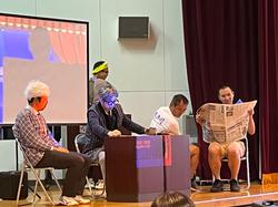 舞台上に置かれた大きなスクリーンの前で、高齢者の扮装をした4人の男性たちが椅子に座っている、寸劇の様子の写真