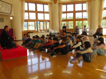 高座に座って話をする落語家の方の講演を床に座って聞いている子供たちを教室の右斜め前から全体を写した写真