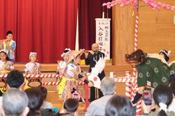 体育館のステージで子どもたちはカラフルな衣装を着て太鼓を演奏しており、獅子舞も登場して演舞が披露されている写真