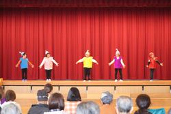 赤い緞帳を背景に、カラフルな衣装を着た5人の子どもたちが整列し、それぞれ帽子をかぶって振り付けに合わせてポーズをとっている写真