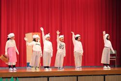 全員が白や淡いピンク系の衣装を着て頭には帽子やカチューシャのような小道具を身に着け6人の子どもたちが並んで手を挙げている劇の写真