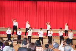 赤い幕を背景に、白いシャツと黒いズボンで揃えた衣装の児童が前後2列に並んで後列の子どもたちは壇上で、手を高く上げたり振り付けをし,前列の子どもたちも拍手をしている発表会での写真