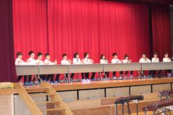 赤い舞台幕の前で全員が白いシャツを着て児童たちが一列に並び観客席に向けた一体感あるパフォーマンスを行っている写真