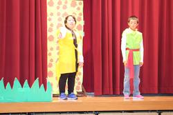 舞台の上で赤いカーテンを背景に、カラフルな衣装を着た二人の子どもが並んで立っている写真