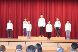 赤いカーテンを背景に、白い服を着た子どもたち6人が舞台の上に整列して立っている写真