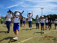 小学校の校庭で行われている運動会で、児童と保護者がバチのような棒を両手に持って、楽しそうに行進している様子の写真