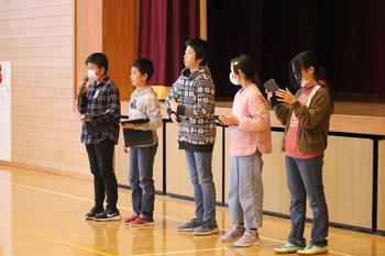 体育館のステージ上で、マイクを持って話している小学生を始め、子どもたちが並んで立っている写真