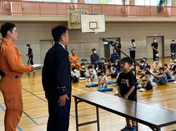 体育館の舞台で長机を挟み消防署の方と1年生の代表が向かい合ってる写真