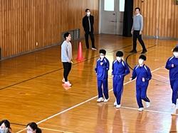 体育館で、体操服姿の生徒たちが講師の男性の指導を受けながら、腕を振っている様子の写真