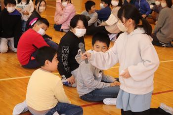体育館の床に座って話を聞いている大勢の小学生たちの中で、マイクを持った女子生徒が男子生徒に話しかけている様子の写真