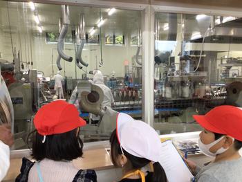 奥に見える工場の機械をガラス越しに見学している子供たちの写真