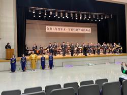 広い舞台の上に警察音楽隊と吹奏楽部の部員たちが楽器を持って並んでおり、その舞台の左下には金色の着物を着た男性を中央に紺色の着物の女性が4人並んで立っている写真