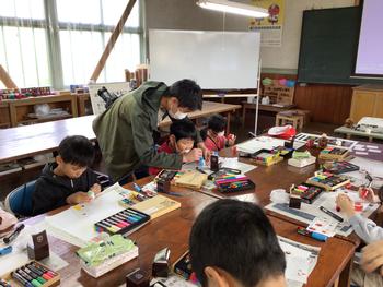 マーカーなどたくさんの道具が置かれた大きな机に向かい合わせに座った複数の児童たちが色付け作業をし、工房の男性が指導をしている様子の写真