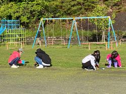 校庭のブランコやすべり台がある遊具のエリアで、数人の保護者や子供たちがしゃがんで、地面の草むしりや清掃活動に取り組んでいる様子の写真