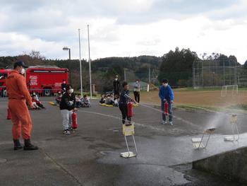 グラウンド横の舗装されたスペースで、消防車が停められ、消防隊員の人が見守る中、3人の児童が訓練用の消火器を使い、的に向かって水をかけている写真